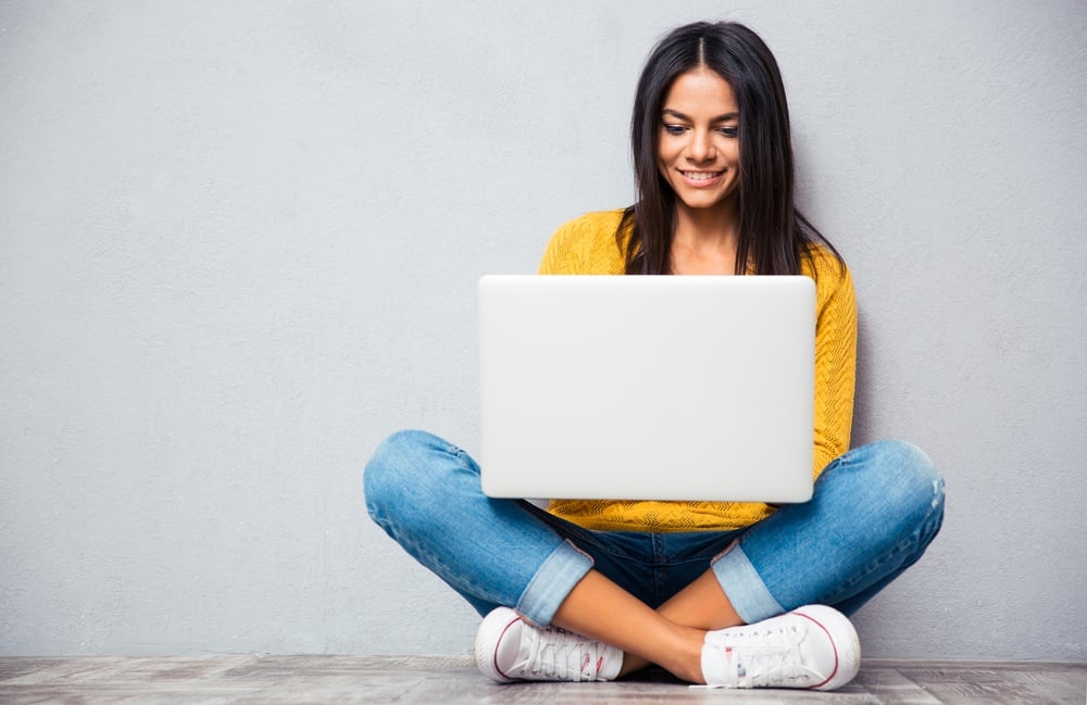 Happy young woman sitting on the floor with crossed legs and using laptop on gray background